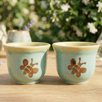 Two small pastel blue-green ceramic cups with a yellow rim and a brown bee design on the front