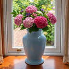 A light blue ceramic vase filled with pink hydrangea flowers and green leaves, placed on a wooden windowsill with a white-framed window behind it.