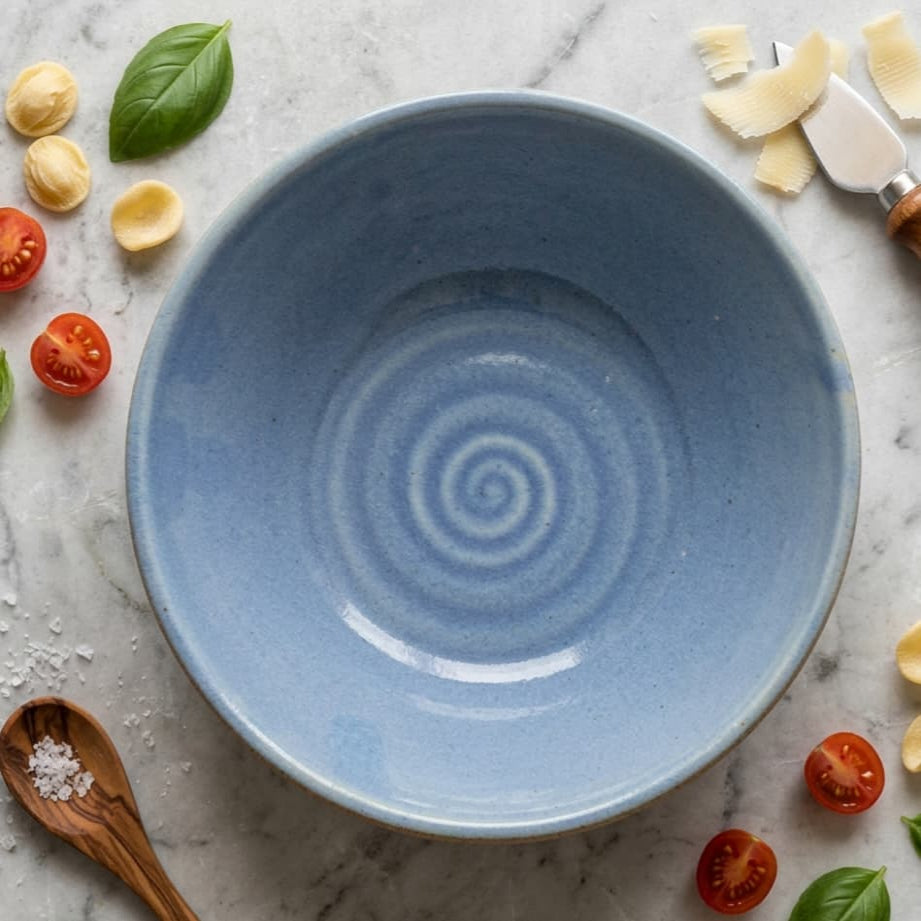 A large round ceramic mixing bowl with a light blue speckled glaze and a spiral pattern in the center, styled on a gray marble surface with pasta ingredients like tomatoes, basil, salt, and pasta around it.