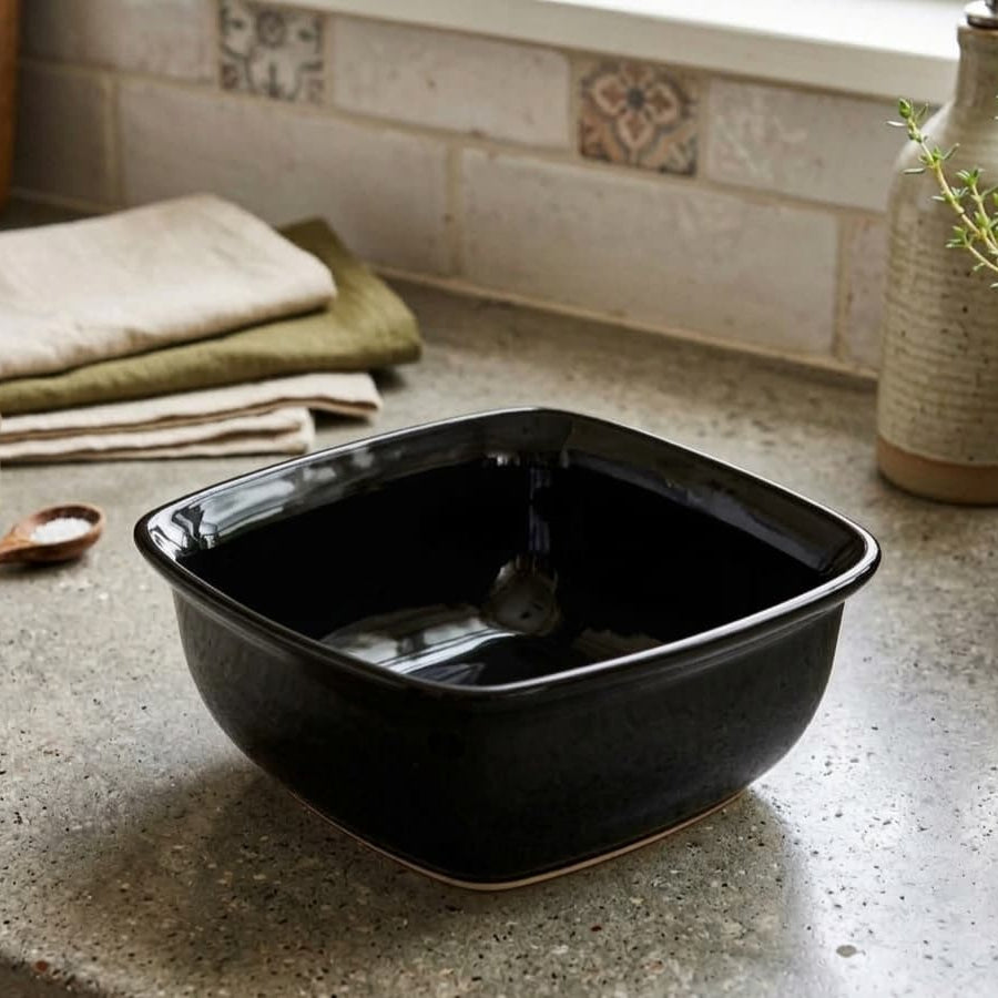 A square black ceramic bowl with a glossy finish, shown on a speckled stone countertop in a kitchen setting with folded towels, a small spoon, and a ceramic vase in the background.