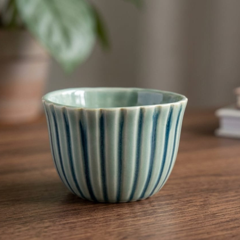 A small ceramic cup with a fluted rim, featuring a pale mint green interior and dark blue-green vertical stripes on an off-white exterior, set on a wooden table with a blurred background.